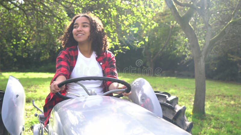 Jeune femme d'adolescente de métis d'afro-américain conduisant un tracteur gris par un champ de pommiers ensoleillé banque de vidéos