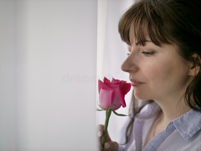 Jeune femme de brune reniflant une rose se tenant prêt la fenêtre image libre de droits Jeune femme de brune reniflant une rose se tenant prêt la fenêtre image libre de droits