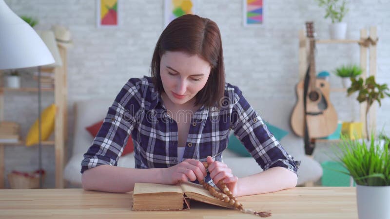 Jeune femme de portrait s'asseyant à une table avec un chapelet dans des ses mains et lisant un livre banque de vidéos