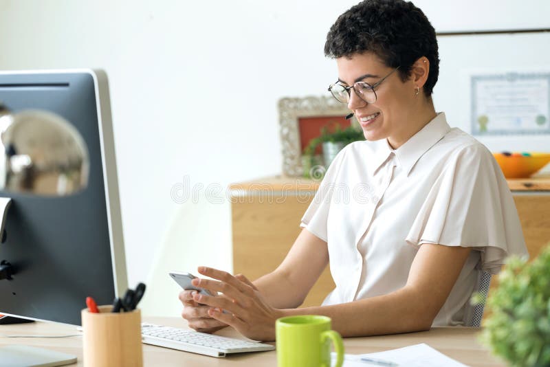 Jeune femme de sourire d'affaires utilisant son smartphone tout en travaillant avec l'ordinateur portable dans le bureau photos stock Jeune femme de sourire d'affaires utilisant son smartphone tout en travaillant avec l'ordinateur portable dans le bureau photos stock