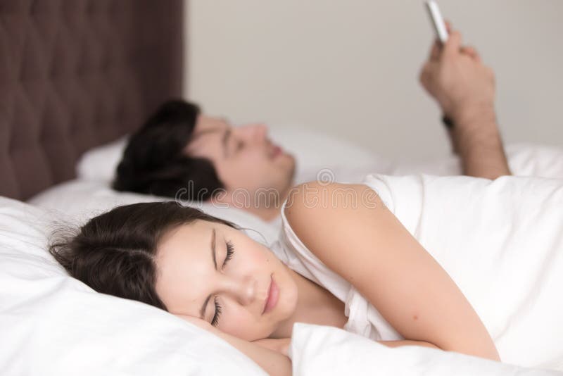 Jeune femme dormant tandis que son ami à l'aide du smartphone dans le lit images stock Jeune femme dormant tandis que son ami à l'aide du smartphone dans le lit images stock