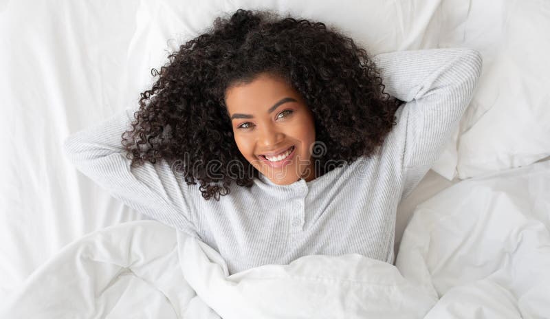 A young Hispanic woman with curly hair is lying on her back in a white bed, resting her head on a pillow. She looks into the camera with a bright, engaging smile. A young Hispanic woman with curly hair is lying on her back in a white bed, resting her head on a pillow. She looks into the camera with a bright, engaging smile