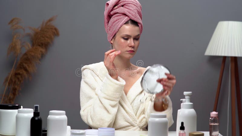 Jeune femme en peignoir avec une serviette sur la tête se regarde dans le miroir et touche le visage sous les yeux après collagène clips vidéos