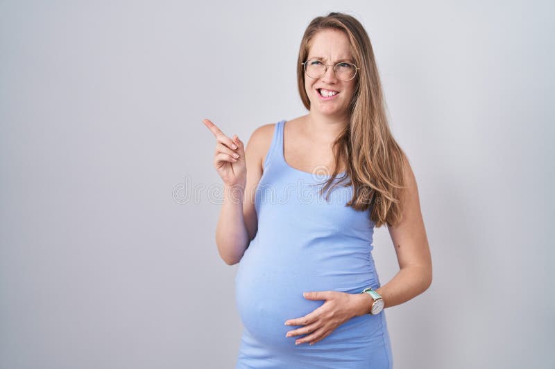 Jeune femme enceinte debout sur fond blanc pointant de côté inquiet et nerveux à l'index concerné et surpris image libre de droits