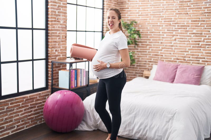 Jeune femme enceinte tenant un tapis de pilate dans la chambre à coucher la langue hors de plaisir avec l'expression drôle photographie stock libre de droits