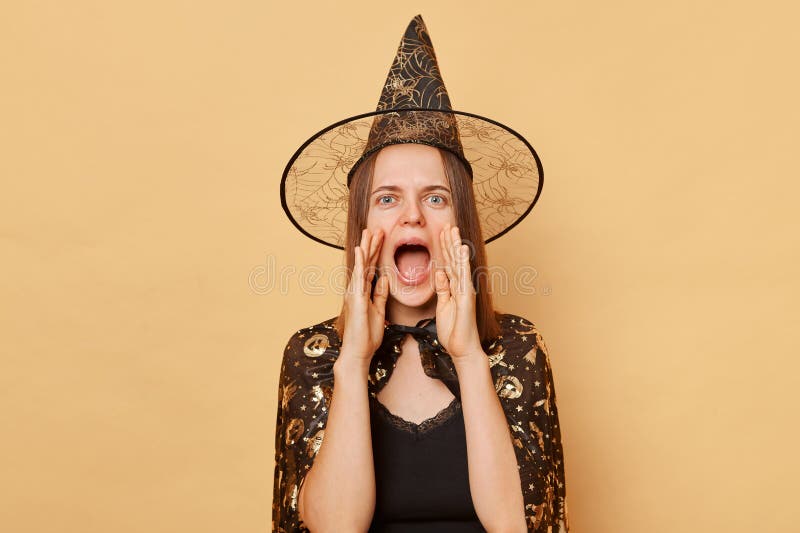 Excited young woman wearing witch costume and carnival cone hat celebrating halloween isolated over beige background making announcement screaming loud with hands near mouth. Excited young woman wearing witch costume and carnival cone hat celebrating halloween isolated over beige background making announcement screaming loud with hands near mouth
