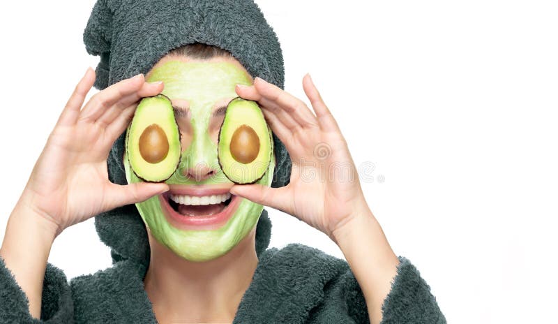 Happy young woman in bathrobe with avocado facial mask applied to her face, smiling and holding avocado fruits cut in half in front of her eyes. Close-up portrait on white background. Happy young woman in bathrobe with avocado facial mask applied to her face, smiling and holding avocado fruits cut in half in front of her eyes. Close-up portrait on white background.