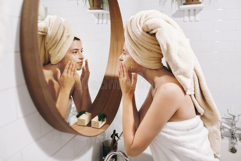 Young happy woman in towel applying organic face mask and looking at round mirror in stylish bathroom. Girl making facial massage with scrub, peeling and cleaning skin on face. Skin Care. Young happy woman in towel applying organic face mask and looking at round mirror in stylish bathroom. Girl making facial massage with scrub, peeling and cleaning skin on face. Skin Care