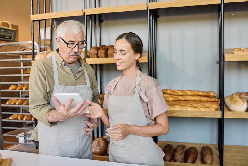 Happy young female baker making presentation of new assortment to mature colleague. Happy young female baker making presentation of new assortment to mature colleague