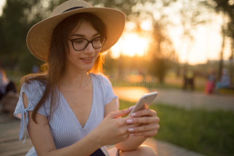 Jeune femme heureuse s'asseyant en parc Souriant et tenant le téléphone dans des ses mains Lumi?re de coucher du soleil image libre de droits Jeune femme heureuse s'asseyant en parc Souriant et tenant le téléphone dans des ses mains Lumi?re de coucher du soleil image libre de droits