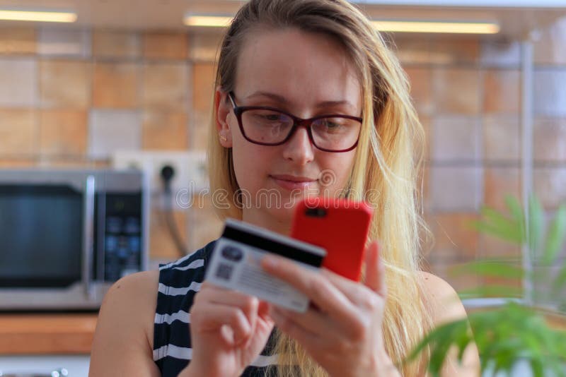 Jeune femme heureuse à l'aide de la carte de crédit et du téléphone dans la cuisine à la maison image stock Jeune femme heureuse à l'aide de la carte de crédit et du téléphone dans la cuisine à la maison image stock