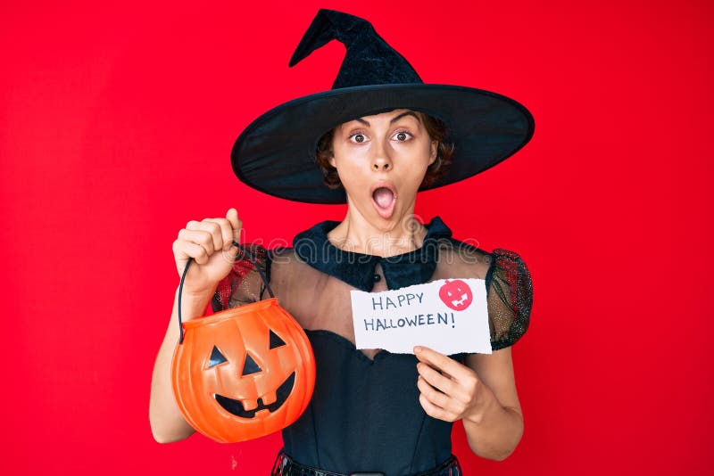 Young hispanic woman wearing witch costume holding pumpkin and happy halloween message afraid and shocked with surprise and amazed expression, fear and excited face. Young hispanic woman wearing witch costume holding pumpkin and happy halloween message afraid and shocked with surprise and amazed expression, fear and excited face