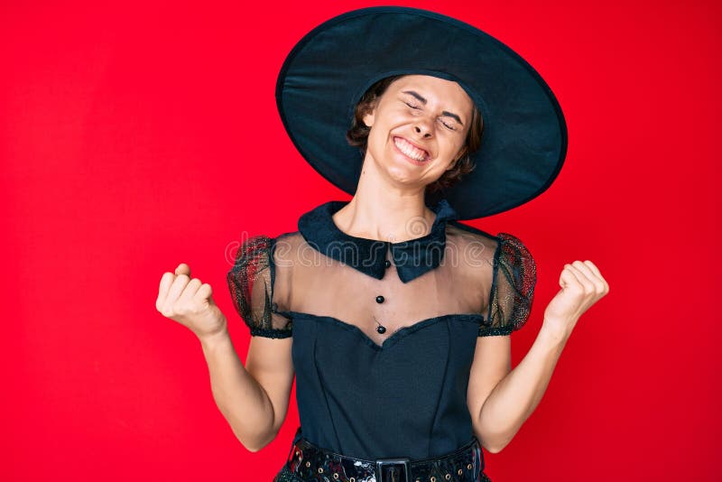 Young hispanic woman wearing witch halloween costume very happy and excited doing winner gesture with arms raised, smiling and screaming for success. celebration concept. Young hispanic woman wearing witch halloween costume very happy and excited doing winner gesture with arms raised, smiling and screaming for success. celebration concept