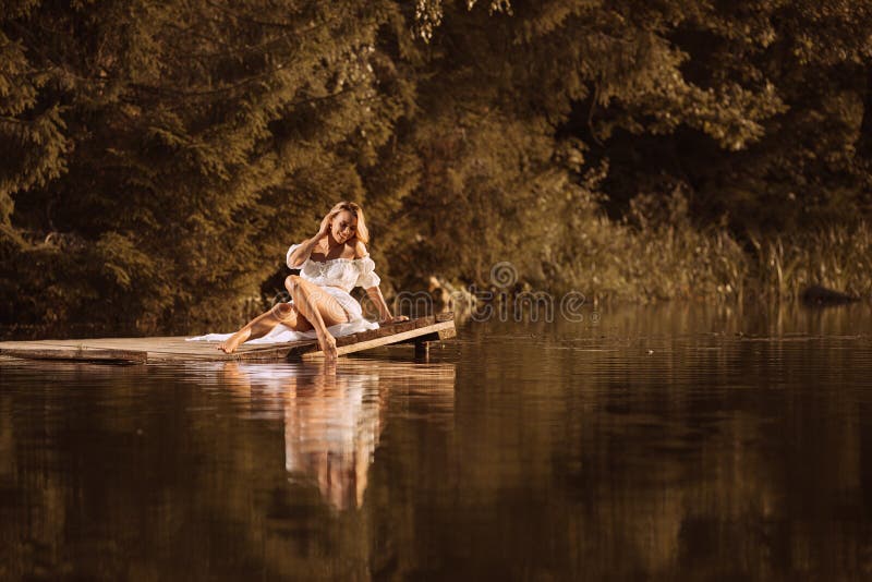 Jeune femme mignonne s'asseyant par le lac touchant l'eau par sa jambe image stock Jeune femme mignonne s'asseyant par le lac touchant l'eau par sa jambe image stock