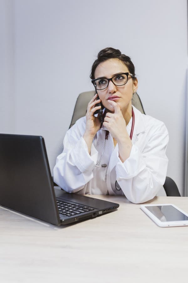 Jeune femme médecin travaillant sur ordinateur portable à la consultation. Parler sur un téléphone portable et avoir l'air inq photos stock Jeune femme médecin travaillant sur ordinateur portable à la consultation. Parler sur un téléphone portable et avoir l'air inq photos stock