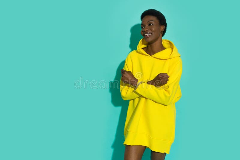 Jeune femme noire souriante dans un capuche jaune vif surdimensionné pose avec les bras croisés et regarde loin photos libres de droits
