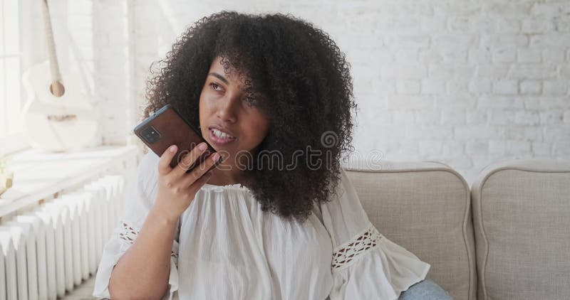 Jeune femme noire souriante enregistrant un message audio à un ami en utilisant un smartphone assis sur un canapé gris banque de vidéos