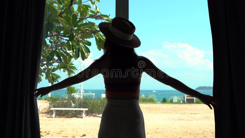 Jeune femme ouvrant les rideaux et voyant la fenêtre vue plage de la mer dans la station balnéaire d'hôtels de luxe banque de vidéos