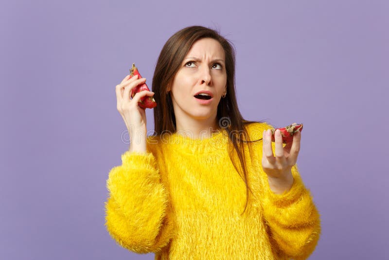 Jeune femme perplexe dans le chandail de fourrure recherchant tenant des halfs du pitahaya, fruit du dragon près des oreilles d'i photos stock Jeune femme perplexe dans le chandail de fourrure recherchant tenant des halfs du pitahaya, fruit du dragon près des oreilles d'i photos stock