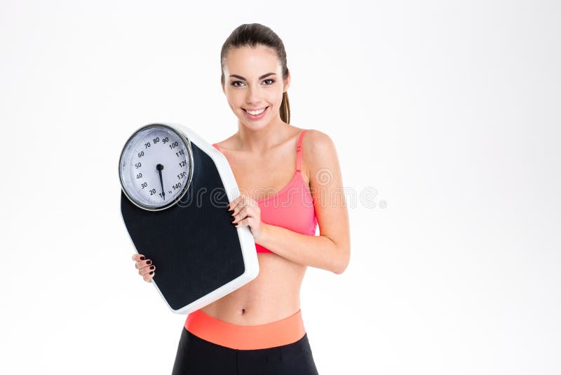 Jeune femme positive de sourire de forme physique dans le sportwear tenant la balance photographie stock libre de droits Jeune femme positive de sourire de forme physique dans le sportwear tenant la balance photographie stock libre de droits