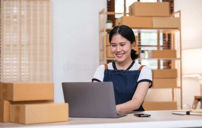 Young woman, small business owner selling products online, accepting online product orders via laptop, sits in a room with boxes of products. Young woman, small business owner selling products online, accepting online product orders via laptop, sits in a room with boxes of products..