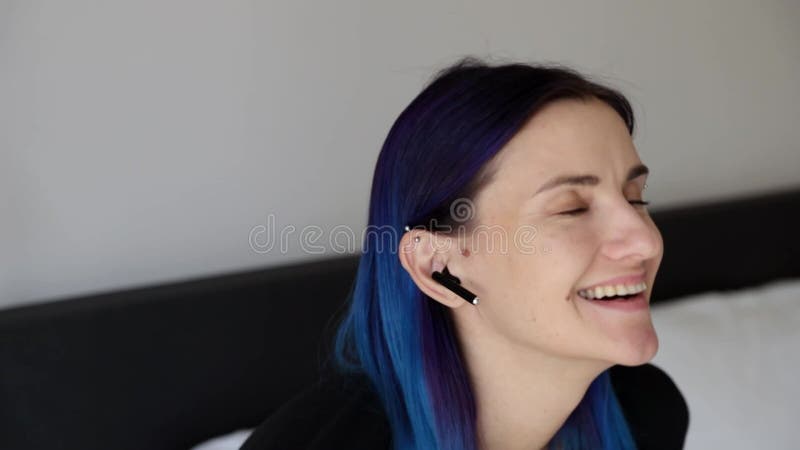 Jeune femme rire bleu de cheveux assis sur le lit avec des écouteurs sans fil banque de vidéos