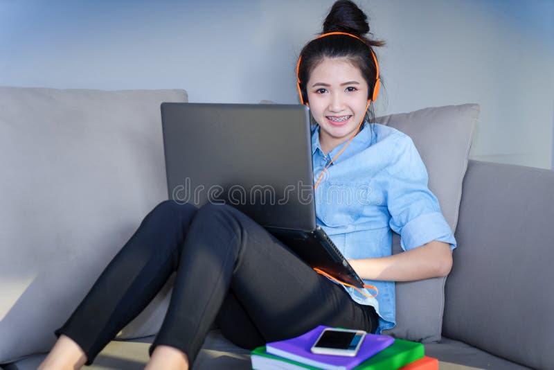Jeune femme s'asseyant sur le sofa avec l'ordinateur portable et l'écouteur liste de fille photographie stock Jeune femme s'asseyant sur le sofa avec l'ordinateur portable et l'écouteur liste de fille photographie stock