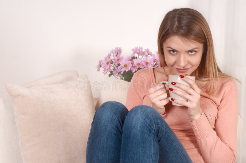 Jeune femme s'asseyant sur le sofa avec la tasse de café ou de thé dans des mains photos stock Jeune femme s'asseyant sur le sofa avec la tasse de café ou de thé dans des mains photos stock