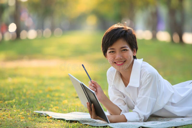 Jeune femme se trouvant sur le parc d'herbe verte avec le crayon et le carnet images libres de droits Jeune femme se trouvant sur le parc d'herbe verte avec le crayon et le carnet images libres de droits