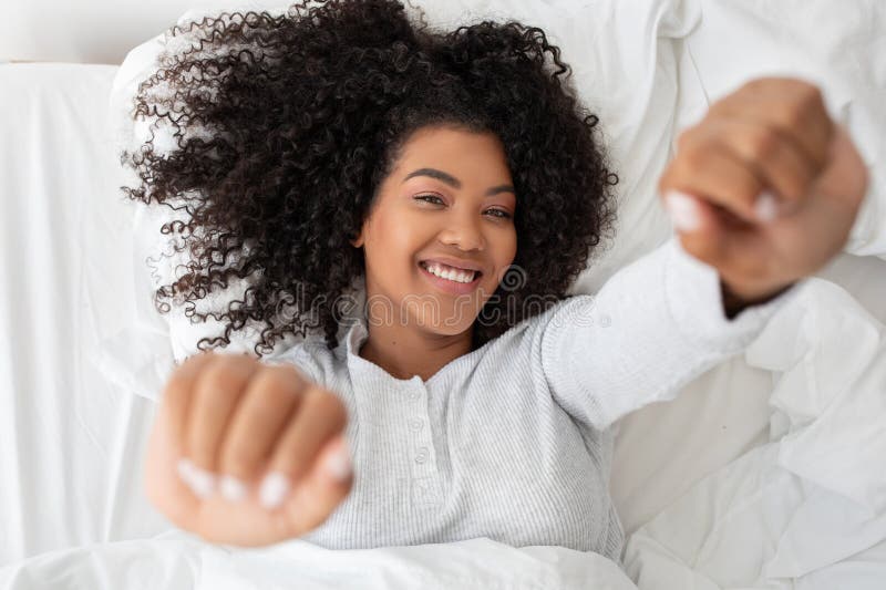 A cheerful young woman with curly hair is lying on her back in a comfy bed, stretching her arms towards the camera with a relaxed and happy expression, suggesting she just woken up. A cheerful young woman with curly hair is lying on her back in a comfy bed, stretching her arms towards the camera with a relaxed and happy expression, suggesting she just woken up.