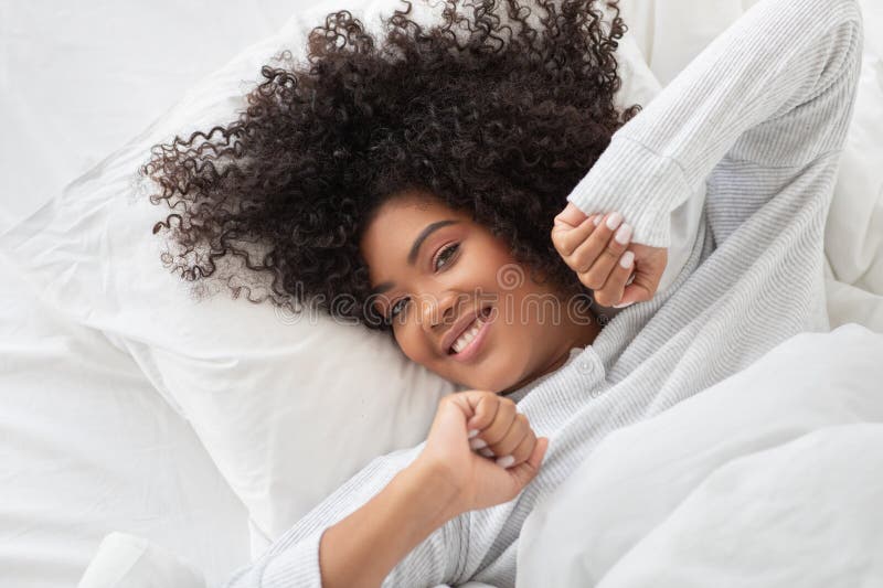 A young Hispanic woman with curly hair is lying in bed, exhibiting a relaxed and happy expression as she gently wakes up. A young Hispanic woman with curly hair is lying in bed, exhibiting a relaxed and happy expression as she gently wakes up.