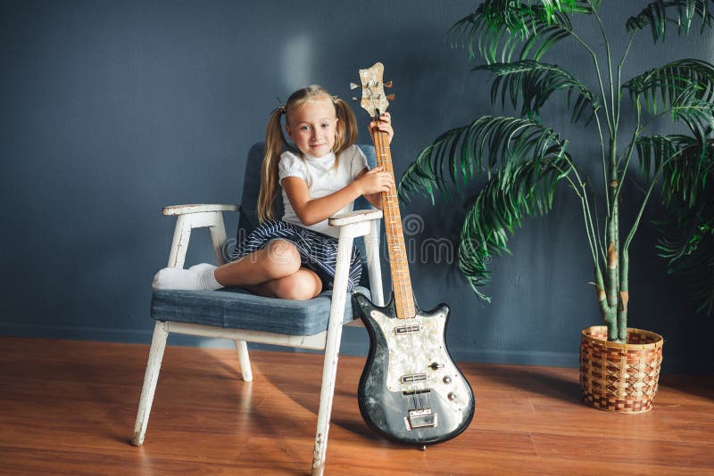 Jeune fille blonde avec des queues en T-shirt, jupe et sandales blancs avec la guitare électrique à la maison regardant la caméra images stock Jeune fille blonde avec des queues en T-shirt, jupe et sandales blancs avec la guitare électrique à la maison regardant la caméra images stock