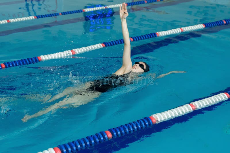 Jeune fille dans les lunettes et chapeau nageant le style arrière de course de rampement dans la piscine d'eau bleue photo stock Jeune fille dans les lunettes et chapeau nageant le style arrière de course de rampement dans la piscine d'eau bleue photo stock