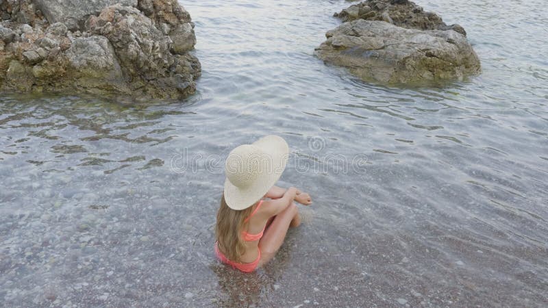 Jeune fille en maillot de bain d'american national standards de chapeau assis dans l'eau de mer sur la plage de galets aux vacanc banque de vidéos