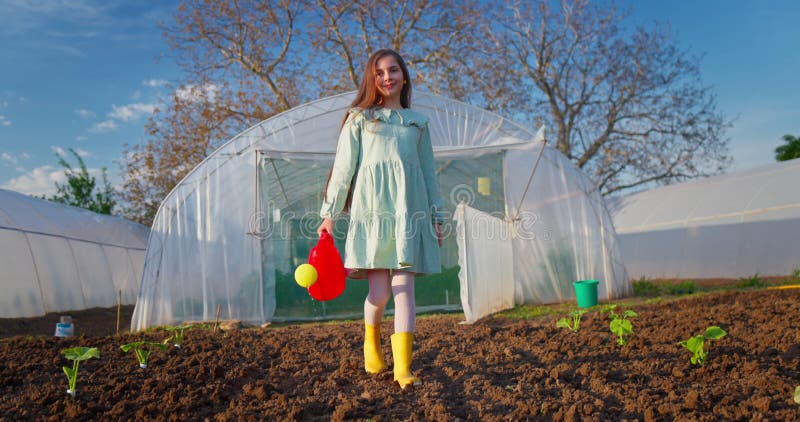 Jeune fille en robe bottes jaunes et arrosage rouge peut marcher dans une serre jardin entouré de jeunes plantes au coucher du sol clips vidéos