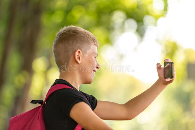 Jeune garçon de la préadolescence heureux marchant dans le jour d'été ensoleillé chaud en parc et prenant le selfie sur le smartp photographie stock libre de droits