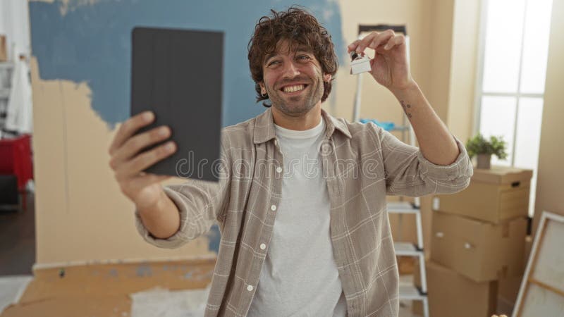 Jeune hispanique souriant lors d'un appel vidéo à la maison tenant les clés et la tablette dans le nouveau salon avec boîtes et éc clips vidéos