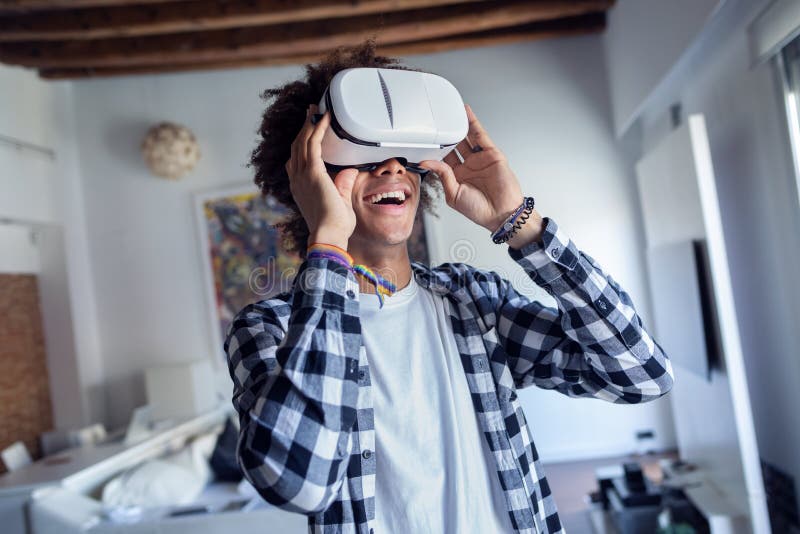 Jeune homme afro-américain heureux employant ses verres de réalité virtuelle tout en se tenant à la maison photo libre de droits Jeune homme afro-américain heureux employant ses verres de réalité virtuelle tout en se tenant à la maison photo libre de droits