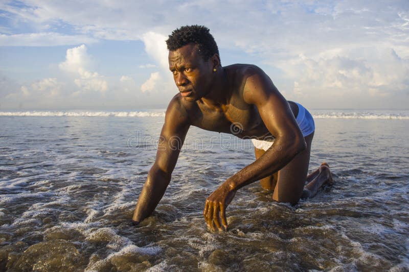 Jeune homme afro-américain noir attirant et sexy avec la pose sportive de corps musculaire fraîche en eau de mer sur la plage de images libres de droits Jeune homme afro-américain noir attirant et sexy avec la pose sportive de corps musculaire fraîche en eau de mer sur la plage de images libres de droits
