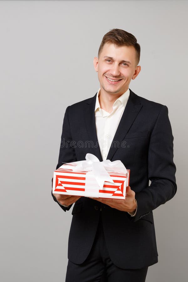 Jeune homme agréable d'affaires dans le costume tenant la boîte actuelle rayée rouge avec le ruban de cadeau d'isolement sur le f image libre de droits Jeune homme agréable d'affaires dans le costume tenant la boîte actuelle rayée rouge avec le ruban de cadeau d'isolement sur le f image libre de droits