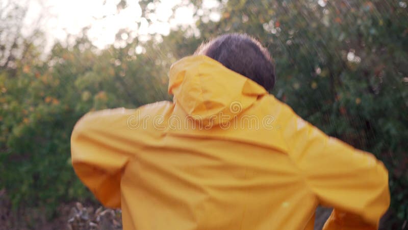Jeune homme arabe souriant portant un imperméable jaune danser sous la pluie dans le parc. mâle joyeux appréciant la pluie banque de vidéos