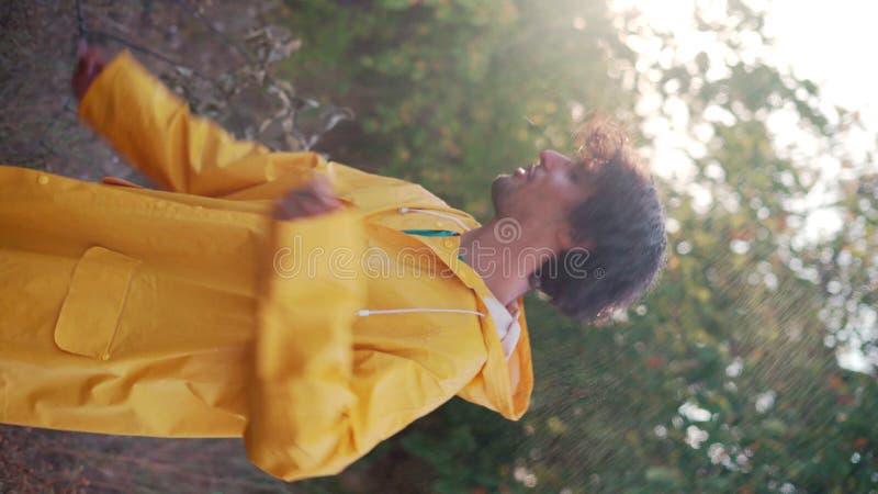 Jeune homme arabe souriant portant un imperméable jaune danser sous la pluie dans le parc. mâle joyeux appréciant la pluie banque de vidéos