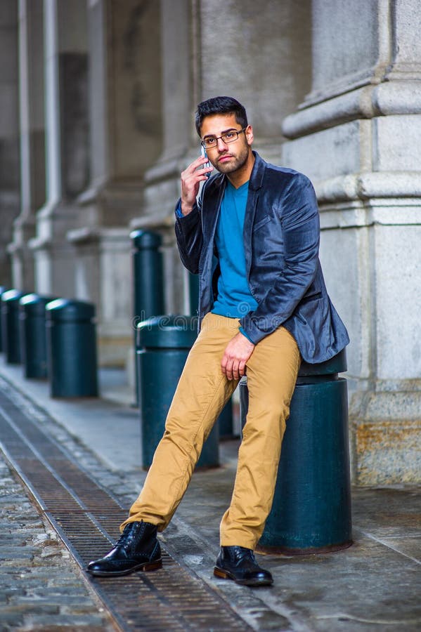 Dressing in  blue woolen blazer, sweater, dark yellow pants and leather boot shoes, wearing glasses, a young guy with bread and mustache is sitting on an old fashion street, making a phone call. Dressing in  blue woolen blazer, sweater, dark yellow pants and leather boot shoes, wearing glasses, a young guy with bread and mustache is sitting on an old fashion street, making a phone call