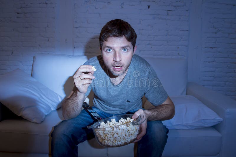 Jeune homme attirant à la maison se trouvant sur le divan regardant la TV tenir le maïs éclaté pour rouler mangeant photographie stock Jeune homme attirant à la maison se trouvant sur le divan regardant la TV tenir le maïs éclaté pour rouler mangeant photographie stock