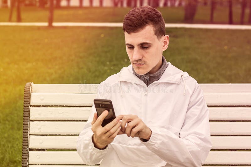 Jeune homme avec le chapeau se reposant sur un banc en bois et vérifiant le message par l'écran de smartphone photographie stock libre de droits Jeune homme avec le chapeau se reposant sur un banc en bois et vérifiant le message par l'écran de smartphone photographie stock libre de droits