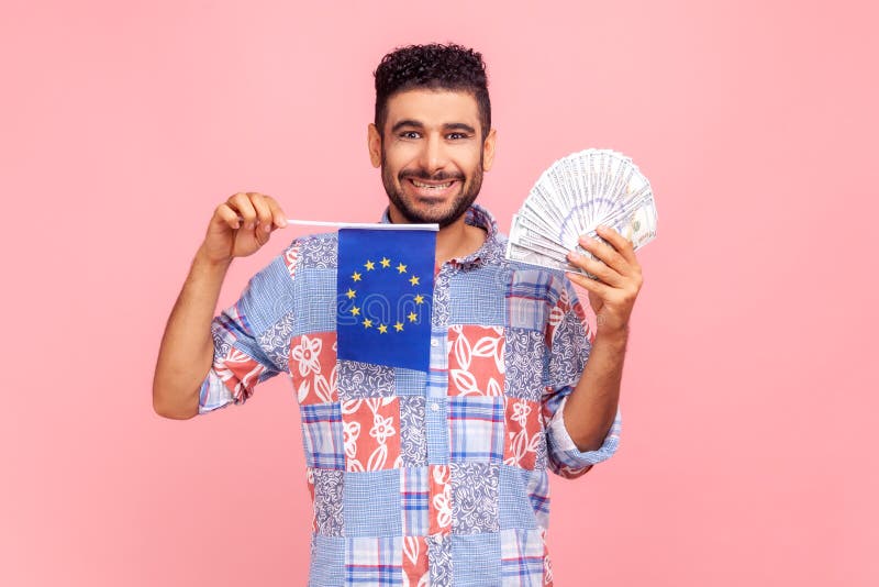 Jeune homme barbu avec un sourire aux dents tenant drapeau européen et des billets regardant la caméra avec une expression faciale image libre de droits