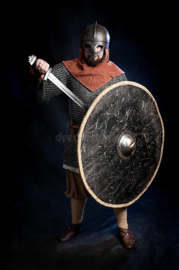 Jeune homme barbu dans un standind et tenir de casque de Viking-ère une épée et un bouclier photo stock Jeune homme barbu dans un standind et tenir de casque de Viking-ère une épée et un bouclier photo stock