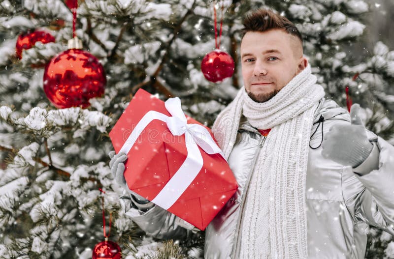 Jeune homme barbu en argent par les détenteurs de manteau à l'arbre de Noël décoré avec le giftbox rouge faisant des gestes comme