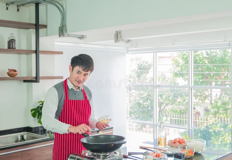 Jeune homme beau et asiatique dans le tablier rouge Cuisson pour la nourriture dans la salle de cuisine à la maison photographie stock libre de droits Jeune homme beau et asiatique dans le tablier rouge Cuisson pour la nourriture dans la salle de cuisine à la maison photographie stock libre de droits