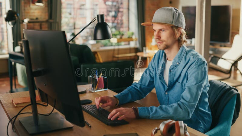 Young Handsome Man Working from Loft Apartment on Desktop Computer. Creative Male Checking Social Media, Browsing Internet from Home. Urban City View from Big Window. Young Handsome Man Working from Loft Apartment on Desktop Computer. Creative Male Checking Social Media, Browsing Internet from Home. Urban City View from Big Window.